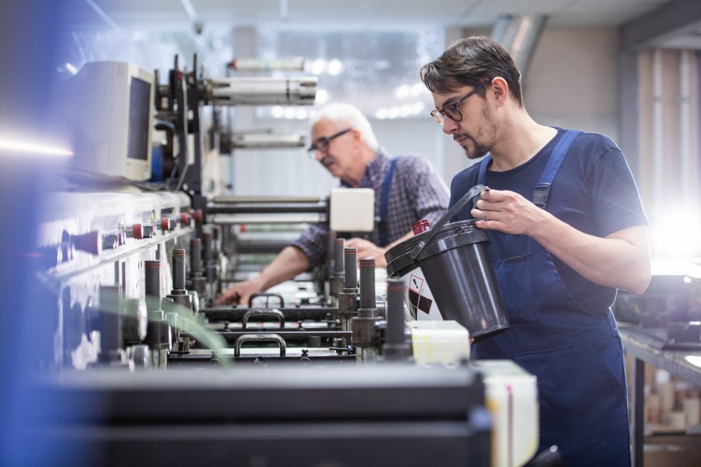 Serious young worker in glasses and overall adding ink into printing press at factory