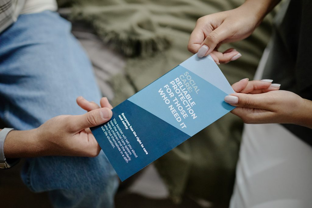 Young adult woman handing informational brochure about social care and protection to teenager during support session, both sitting closely together
